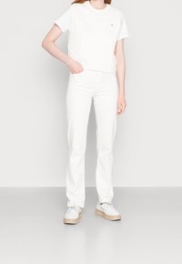 Vit stickad väst med kabelmönster över en vit t-shirt, i kombination med vita raka jeans och ljusa sneakers med tjock sula.