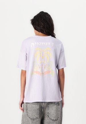 Persona que lleva una camiseta de color púrpura claro de O’Neill Surf Co. con un gráfico tropical desvaído en amarillo y naranja en la parte trasera y vaqueros de mezclilla grises.