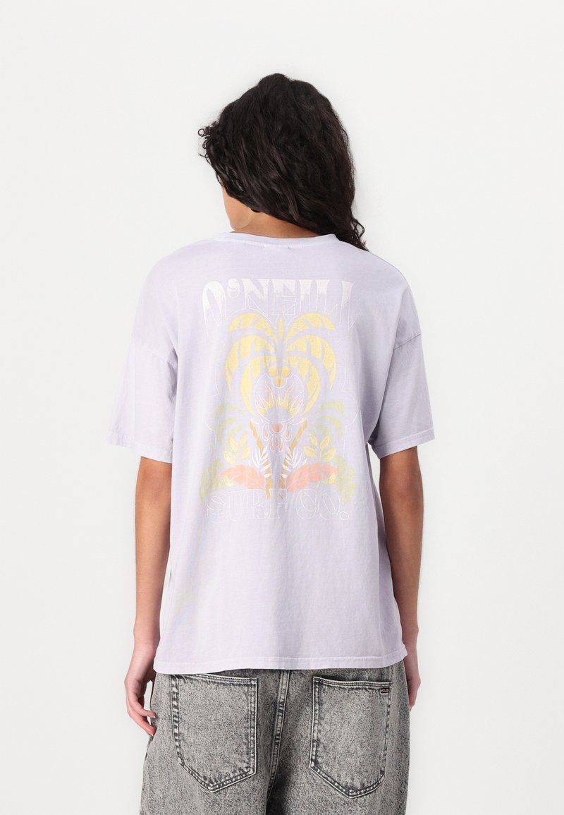 Personne portant un t-shirt violet clair O’Neill Surf Co. avec un motif tropical jaune et orange délavé dans le dos et un jean en denim gris.