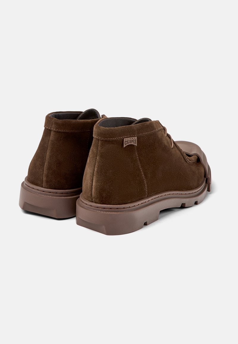 Braune Wildleder-Stiefeletten mit robustem Gummisohle, einfache Schnürung und seitlichem Etikett mit Markenname. Glatte Textur, abgerundete Form.