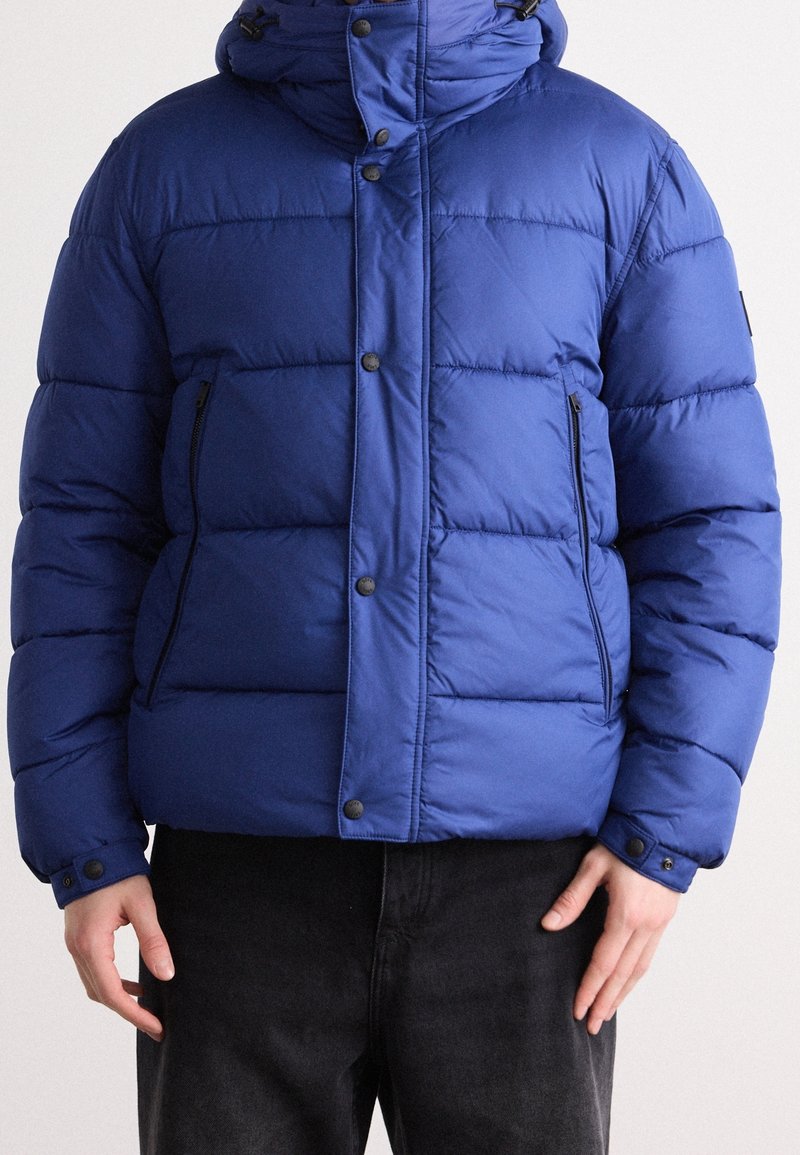 Homme portant une veste d'hiver bleue matelassée avec fermeture éclair et boutons-pression, debout les mains relâchées le long du corps sur un fond uni.
