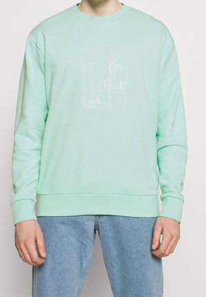 Mann trägt ein hellgrünes Sweatshirt mit einer weißen minimalistischen Linienzeichnung eines Gebäudes und einer Palme auf der Vorderseite, kombiniert mit blauer Jeans.