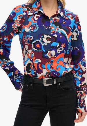 Langærmet skjorte med knapper og kraftige blomster- og abstrakte mønstre i blå, lilla, orange og hvid, kombineret med sorte jeans og sort bælte.