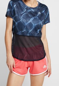 Kobieta w niebieskiej koszulce Adidas z wzorem i czarną siateczką oraz koralowych szortach Adidas z białymi wykończeniami na tle jednolitego tła.