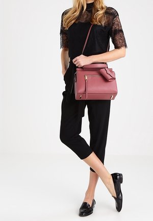 A woman in a black lace top and cropped black pants holds a pink handbag, standing against a light background.