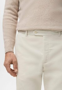 Suéter beige de canalé y pantalones crema claro de canalé con cinturón a juego y detalle de botones, mostrando una tela texturizada. Mano apoyada sobre los pantalones.