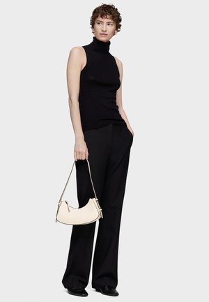 Woman with short curly hair wearing a black sleeveless turtleneck and wide black pants, holding a cream handbag in her left hand.