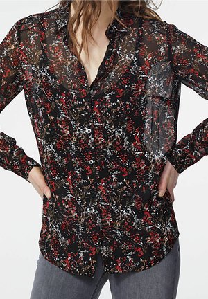 Femme portant un chemisier noir transparent boutonné avec des motifs floraux rouges, blancs et bruns, associé à un pantalon gris, mains sur les hanches.
