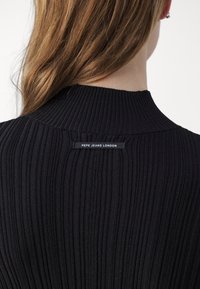 Femme aux longs cheveux bruns portant un pull noir côtelé avec l'étiquette "Pepe Jeans London" centrée dans le haut du dos.