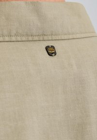 Lys beige linned skjorte med krave og en lille sort og guld mærkat på bagsiden, der fremhæver enkel søm og let tekstur.