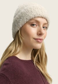Gorro tipo beanie en color crema, hecho de un material suave y texturizado. Forma redonda, con el borde ligeramente rizado. Llevado con un suéter de color burdeos oscuro.