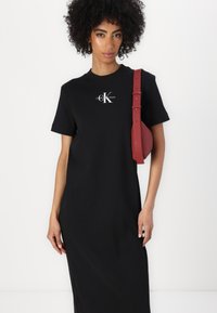 Juodas medvilninis t-shirt su trumpomis rankovėmis, su baltu Calvin Klein logo. Suderintas su raudonu įstrižiniu maišu, kuriam būdinga įgaubta forma.