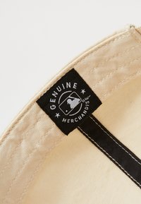 La tela beige con costuras negras presenta una etiqueta negra que dice "MERCANCÍA GENUINA" y un logotipo de béisbol, cosido dentro de la banda del sombrero.