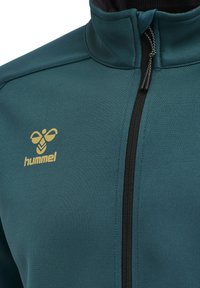 Tijgerwaarzjacket in teal met hoge kraag, voorzien van een gouden logo op de linkerborst en een zwarte ritssluiting. Glad textuur, atletisch ontwerp.