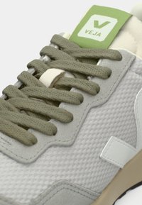 Ανοιχτόγκρι αθλητικά παπούτσια με επάνω μέρος από υφή πλέγματος, κορδόνια σε ελαιόχρωμο πράσινο και ετικέτα λογότυπου σε πράσινο χρώμα· λαστιχένια σόλα με αντίθετο σχέδιο.