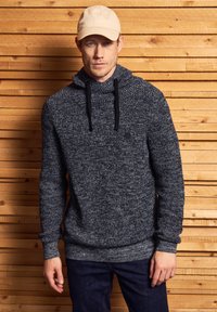 Mörkgrå hoodie med en strukturerad stickning, med en krage, svarta dragsnören och en cirkulär logotypdetalj. I kombination med mörka jeans.