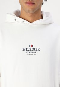 Bílá mikina s kapucí a šňůrkou, na přední straně s logem, které zní "HILFIGER NEW YORK ZALOŽENO 1985" s červenými a modrými akcenty.