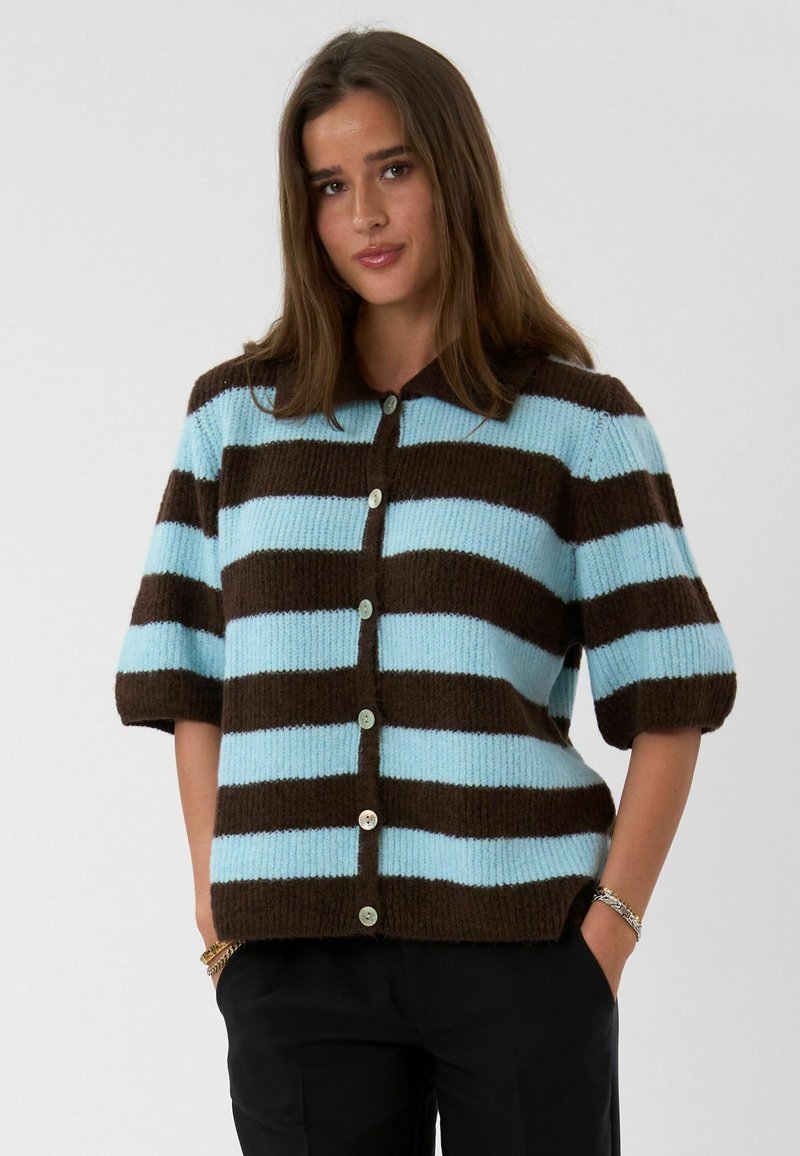 Femme portant un gilet tricoté à manches courtes boutonné avec de larges rayures horizontales marron et bleu clair, debout les mains dans les poches.