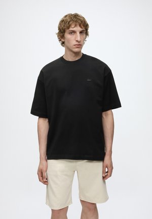 Jeune homme aux cheveux bouclés portant un t-shirt noir ample et un short beige clair, debout devant un fond blanc uni.