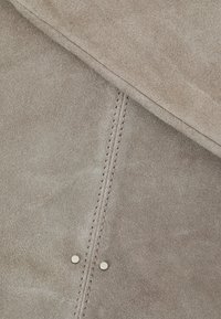 Beige mockaläder med en slät yta, prydd med subtila sömmar och två metallknappar längs sömmen för extra detalj.