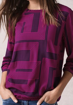 Femme portant un haut violet à manches longues avec un motif géométrique en lignes rose vif, mains dans les poches, et cheveux longs bruns partiellement visibles.