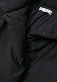 Veste matelassée noire avec des boutons-pression et une étiquette de marque blanche à l'intérieur du col, présentée partiellement ouverte.