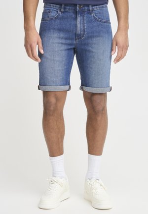 Denim shorts i en medium blå vask med opfoldede kanter, fem lommer og en klassisk pasform, parret med hvide sneakers og sokker.