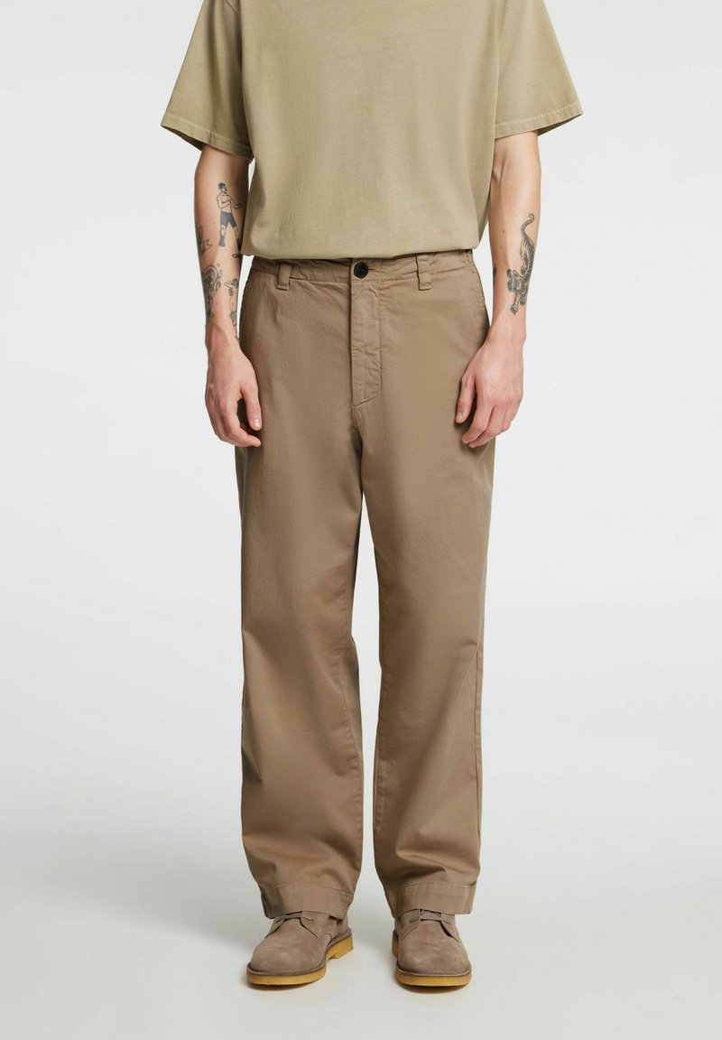 Personne portant un pantalon large beige, un t-shirt beige assorti et des chaussures marron, avec des tatouages visibles sur les deux avant-bras.