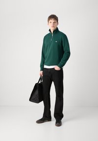 Grønn zip-neck hettegenser, hvit skjorte under, sorte bukser og mørke joggesko, holder en svart bag med teksturet mønster.