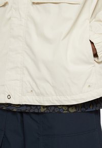 Světlý béžový kabát s hladkou texturou, vybavený kapsou a zapínáním na patentky. Pod ním je vrstva s tmavě modrým vzorem a zelenými a krémovými akcenty.