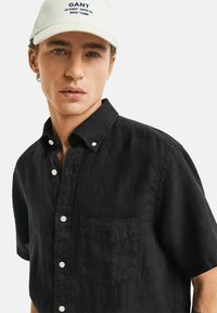 Schwarzes Kurzarm-Hemd mit Knöpfen und spitzem Kragen, mit einer Brusttasche, weißen Knöpfen und strukturiertem Stoff. Vor einem schlichten Hintergrund aufgenommen.