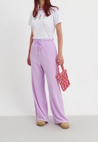 Pantalons larges lilas en tissu texturé avec taille à cordon de serrage, associés à un t-shirt blanc orné de broderies florales. Tenant un petit sac tissé multicolore.