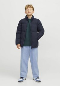Marineblaue Steppjacke mit hohem Kragen, gestepptem Design und seitlichen Taschen. Kombiniert mit einem dunkelgrünen Pullover und hellblauen, weit geschnittenen Jeans.