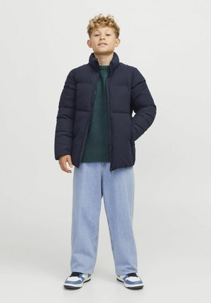 Doudoune marine avec col haut, design matelassé et poches avant. Associée à un pullover vert foncé et un jean large bleu clair.