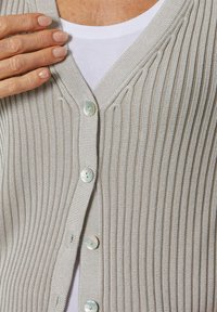 Helles graues, geripptes Cardigan mit tiefem V-Ausschnitt, verziert mit perlenähnlichen Knöpfen und einer sanften Textur, über einem weißen Hemd getragen.