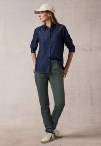 Camisa de botón de manga larga color azul oscuro; pantalones verdes olivo; zapatillas blancas; gorra beige; mujer posando frente a un fondo neutro.