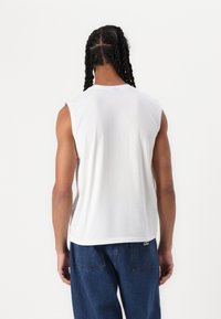 Canottiera bianca senza maniche in cotone, con scollatura rotonda, vestibilità comoda e un leggero logo vicino all'orlo, abbinata a jeans in denim blu.