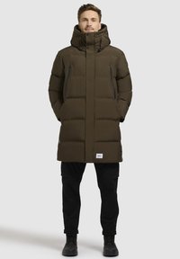 Sötétzöld puffertakaró magas gallérral és állítható kapucnival. Két cipzáras zsebe van, és a szegélyen egy címke található. Az anyag simának és melegnek tűnik.