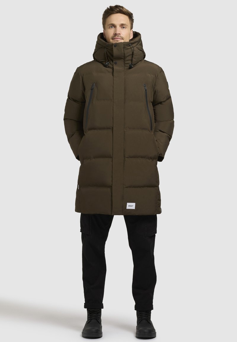 Sötétzöld puffertakaró magas gallérral és állítható kapucnival. Két cipzáras zsebe van, és a szegélyen egy címke található. Az anyag simának és melegnek tűnik.