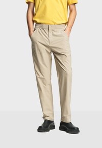 Homme portant un pantalon beige avec taille élastique et bouton, t-shirt jaune, bottes noires, mains dans les poches, debout devant un fond uni.