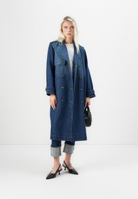 Lang blå denimkåpe med dobbeltspent design, stor krage og gullknapper. Kombinert med oppbrettede jeans og svarte hælssko.