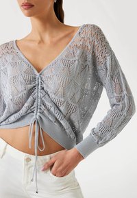 Light blue knitted sweater with a V-neck, long sleeves, and gathered front detail; features a textured leaf pattern and ribbed cuffs.