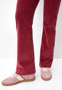 Rode corduroy flared broek gecombineerd met roze suède sneakers met witte accenten en veters, tegen een neutrale achtergrond.
