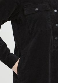 Cămașă neagră din catifea cu nasturi în față, două buzunare la piept și mâneci lungi. Prezintă un material texturat și un ușor luciu.