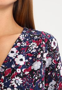 Blomsterblus med en marinblå bakgrund och rosa samt vita blommor, V-hals och rynkdetaljer vid halslinningen.