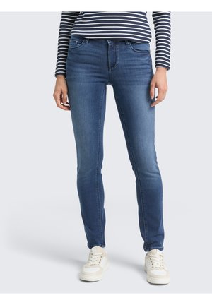 Personne portant un jean bleu slim, un haut à manches longues rayé et des baskets blanches, debout devant un fond clair uni.