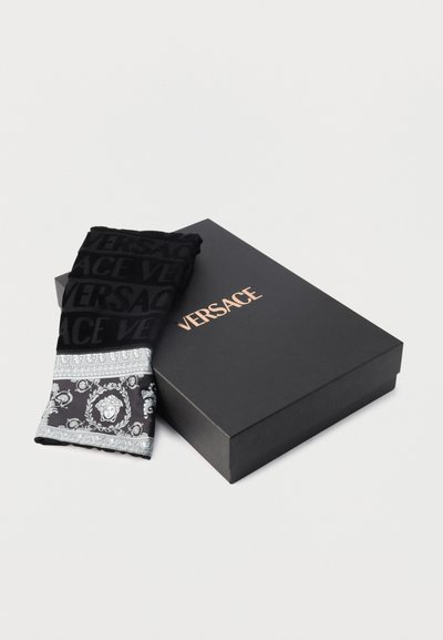 Μαύρο κασκόλ Versace με λογότυπο και σχέδιο Μέδουσας δίπλα σε μια μαύρη συσκευασία Versace πάνω σε λευκό φόντο.