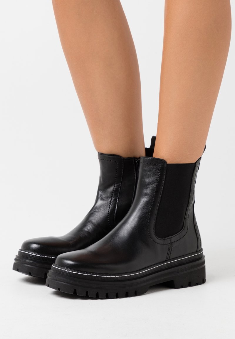 Bottes cheville en cuir noir avec semelles crantées épaisses portées sur des jambes nues sur un fond blanc.