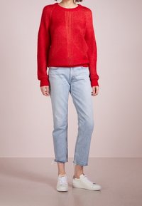 Röd stickad tröja med ett strukturerat mönster, rund halsringning och ribbade manschetter, kombinerad med ljusblå raka jeans och vita sneakers.