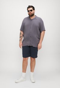 Camisa de manga corta y botones en gris claro, combinada con pantalones cortos a rayas en azul marino. El modelo lleva zapatillas blancas y gafas de sol. Fondo liso.